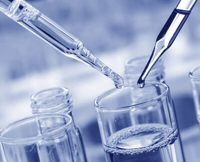 Two pipettes are positioned above a glass container filled with liquid, with droplets poised to fall into it. The image has a blue tint, creating a laboratory atmosphere.