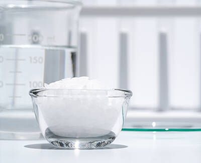 A clear glass bowl filled with white granules sits in front of a measuring beaker containing water, set against a bright, reflective surface.