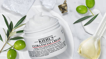A jar of Kiehl's Ultra Facial Cream surrounded by olive branches, a small bowl of cream, and a decorative spoon on a marble surface.