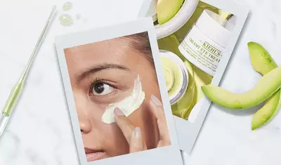 A close-up of a person applying a creamy eye treatment under their eye, with jars of the product and slices of a green fruit in the background. The image is styled like a polaroid photo.