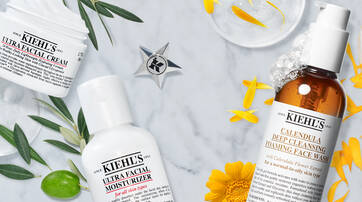 A marble surface featuring Kiehl's skincare products, including Ultra Facial Cream, Ultra Facial Moisturizer, and Calendula Deep Cleansing Foaming Face Wash, surrounded by yellow flowers and green leaves.