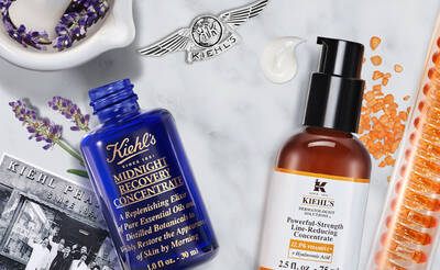 Flat lay of Kiehl's skincare products, including a blue bottle labeled "Midnight Recovery Concentrate" and an orange bottle labeled "Powerful-Strength Line-Reducing Concentrate," surrounded by lavender, orange capsules, and a vintage Kiehl's product card against a marble background.