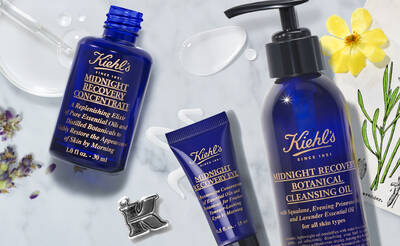 Alt text: A flat lay of Kiehl's Midnight Recovery skincare products, including a blue bottle of recovery concentrate, a tube of recovery eye treatment, and a cleansing oil bottle, surrounded by botanical elements and a flower.