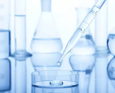A clear pipette is poised above a petri dish with a droplet about to fall, surrounded by laboratory glassware in shades of blue.