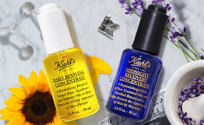 Two Kiehl's skincare products are displayed: a yellow bottle labeled "Daily Reviving Concentrate" and a blue bottle labeled "Midnight Recovery Concentrate." Both are placed among sunflowers, lavender, and a scientific glass element, highlighting their natural and restorative properties.