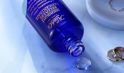 A blue bottle of Tatcha's Midnight Concentrate skincare product, partially spilled on a marble surface, with a crystal-like droplet nearby and some gold rings in the background.