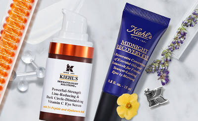 A flat lay of Kiehl's skincare products, featuring a pump bottle of Powerful-Strength Line-Reducing & Dark Circle-Diminishing Vitamin C Eye Serum and a tube of Midnight Recovery Eye cream, surrounded by orange capsules, lavender, and a flower.