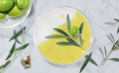 A clear bowl filled with a smooth, green mixture sits on a marble surface, accompanied by fresh green olives and olive leaves. A small mortar and pestle is placed nearby, suggesting an olive oil preparation theme.