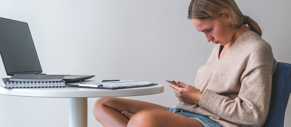 Młoda dziewczyna siedzi przy komputerze, ale patrzy w smartfona, garbiąc się i przyjmując postawę tech neck