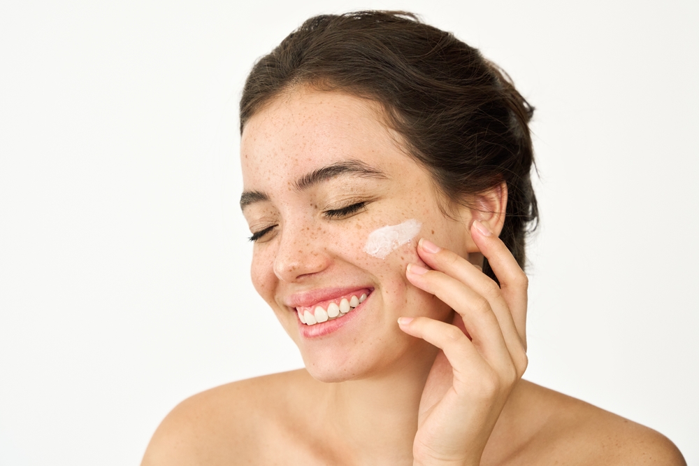 Uśmiechnięta młoda dziewczyna nakłada krem przeciwzmarszczkowy na twarz i pod oczy